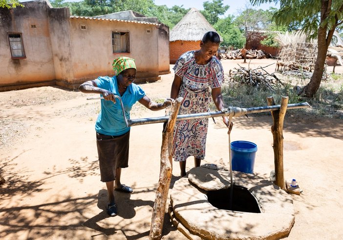 Faith Chasakara and her friend collect water in the well in her village, near Bindura in Zimbabwe. Faith is wearing a pretty patterned dress, a blue bucket sits to the side of the well and there are homes in the background with trees dotted around.