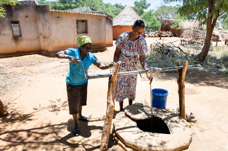 Faith Chasakara and her friend collect water in the well in her village, near Bindura in Zimbabwe. Faith is wearing a pretty patterned dress, a blue bucket sits to the side of the well and there are homes in the background with trees dotted around.