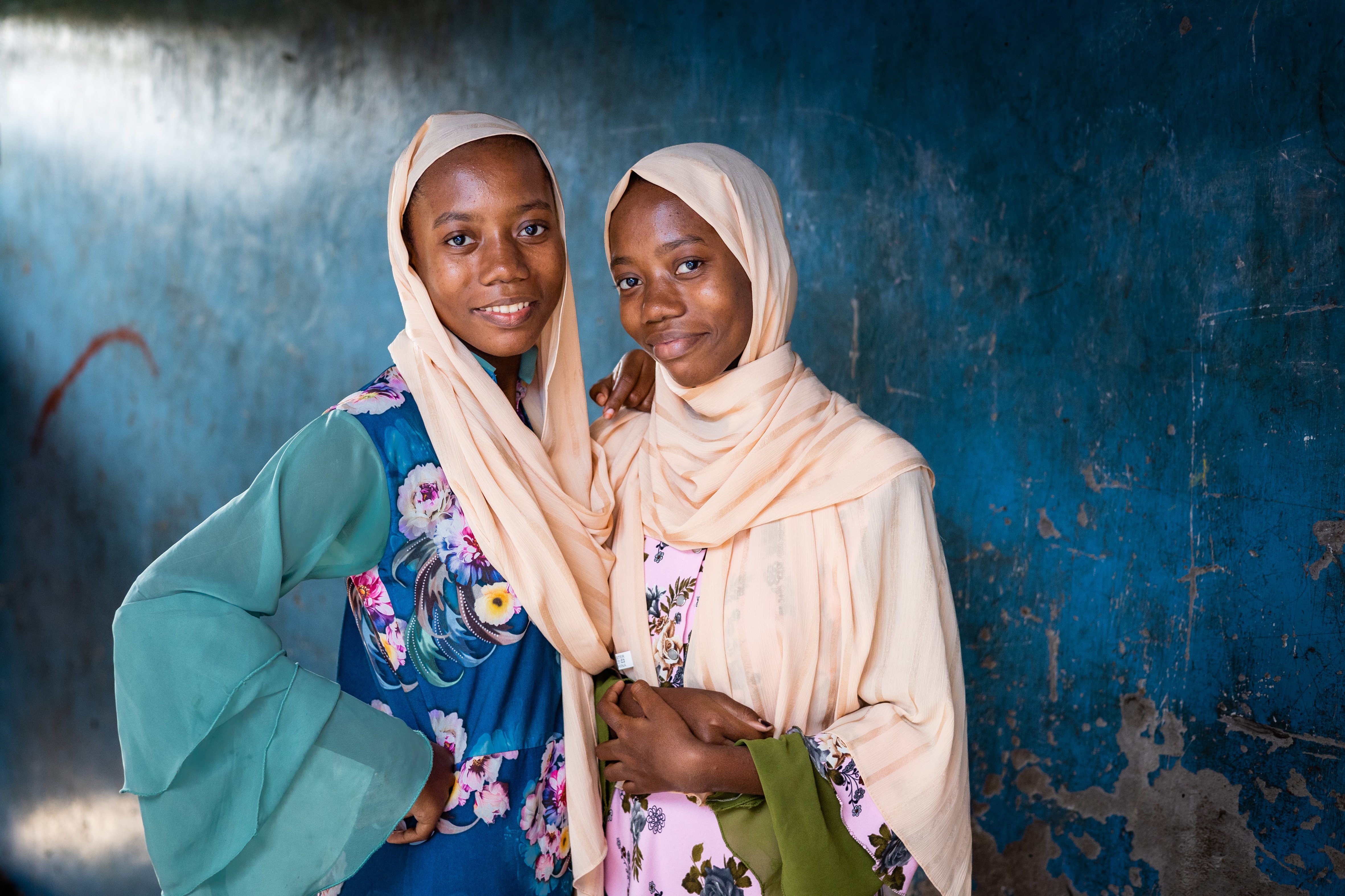 Two girls stand in front of a blue wall smiling to camera